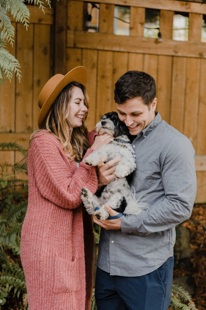 Middle aged man and woman laughing and holding black and white biewer terrier dog in front of orange painted fence.