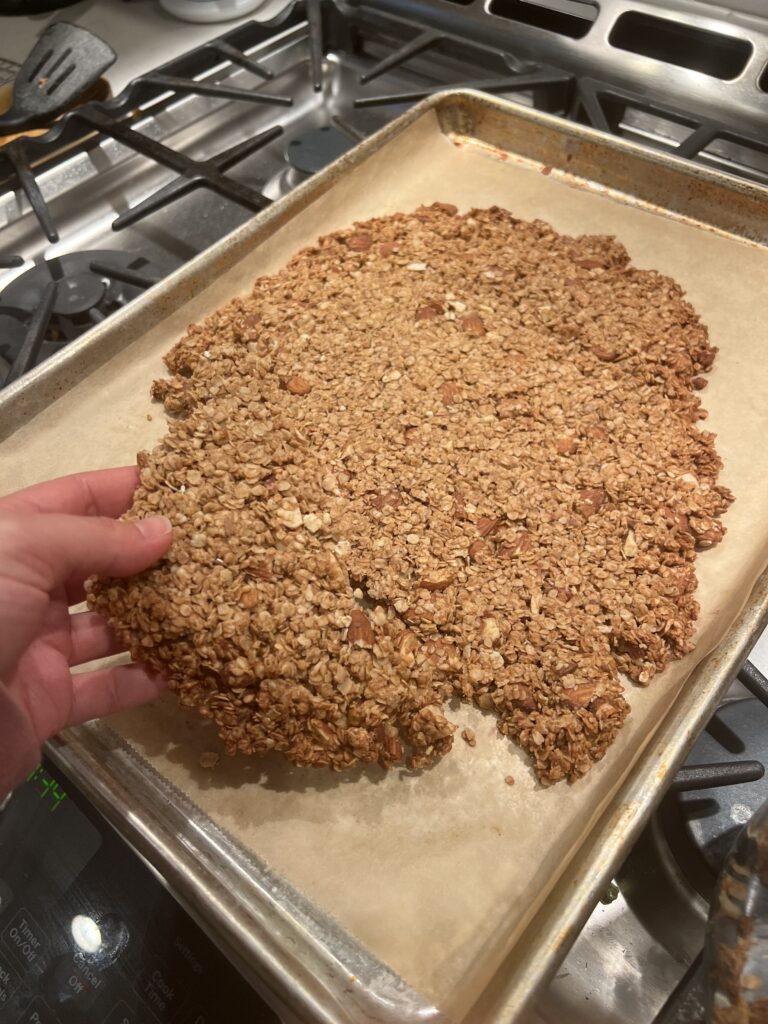 Hand holding a piece of Honey Almond granola  from a lined baking sheet on top of the stove