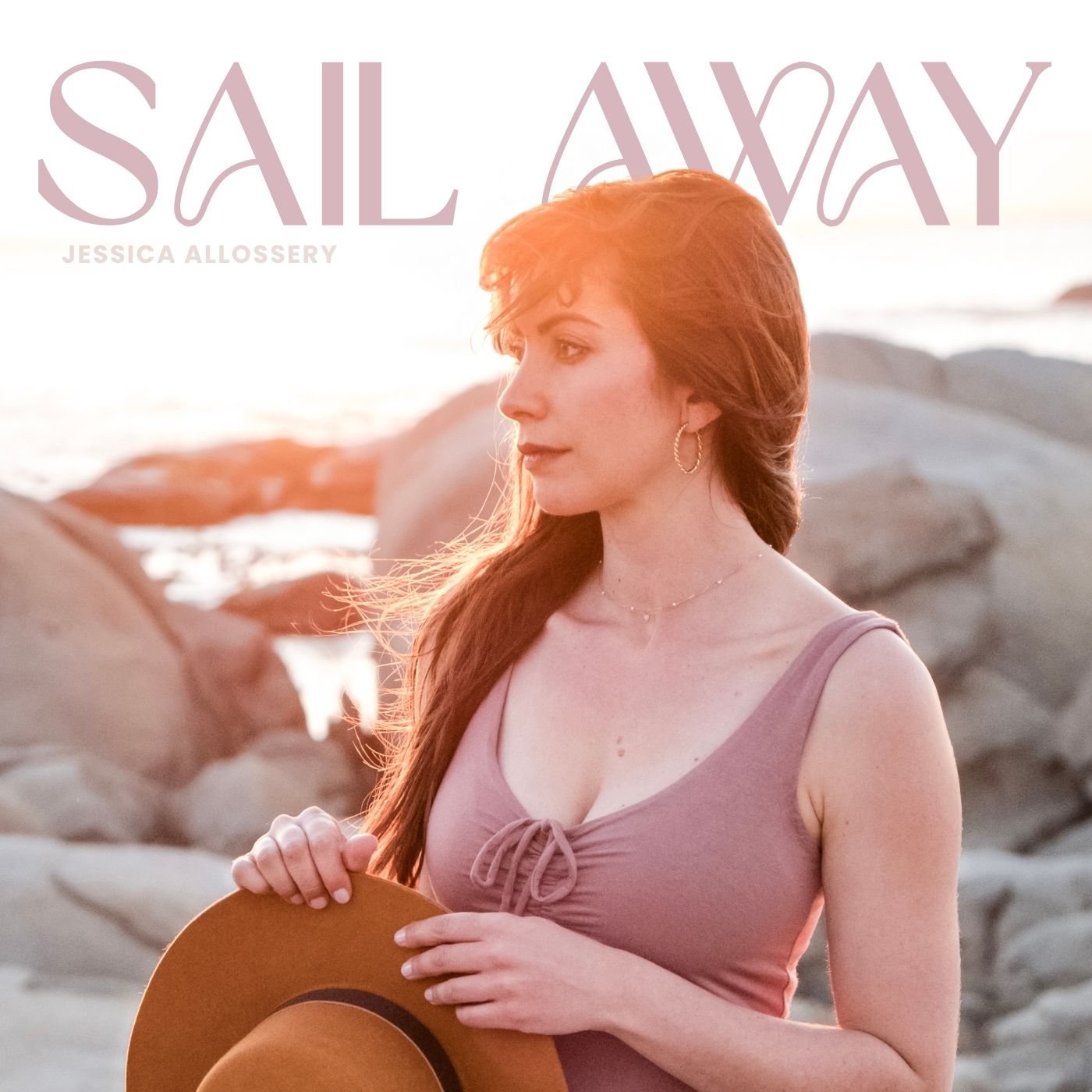 photo of a woman standing in front of a rocky shore at sunset, holding a wide brimmed hat wearing a pink dress
