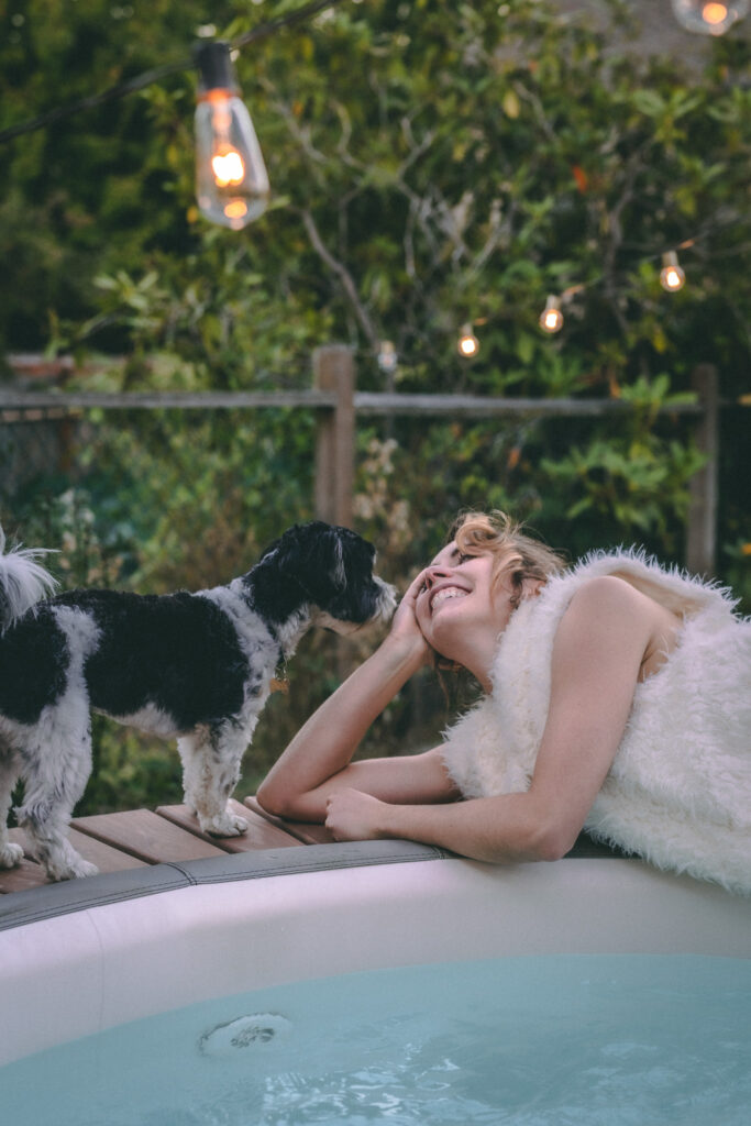 Woman smiling at black and white biewer terrier dog laying on edge of blow up hot tub in PNW yard.