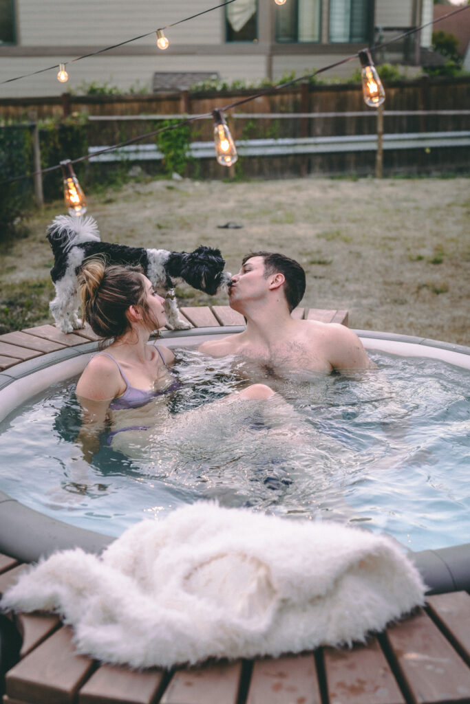 Middle aged Woman and man in hot tub, with black and white biewer terrier dog giving kiss to mans face on edge of blow up hot tub in yard.